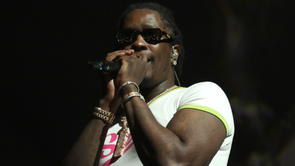 A performer wearing sunglasses and a white shirt sings into a microphone on stage.