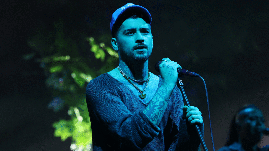 A man with tattoos and a blue cap sings into a microphone on stage, under blue lighting, with a blurred background.