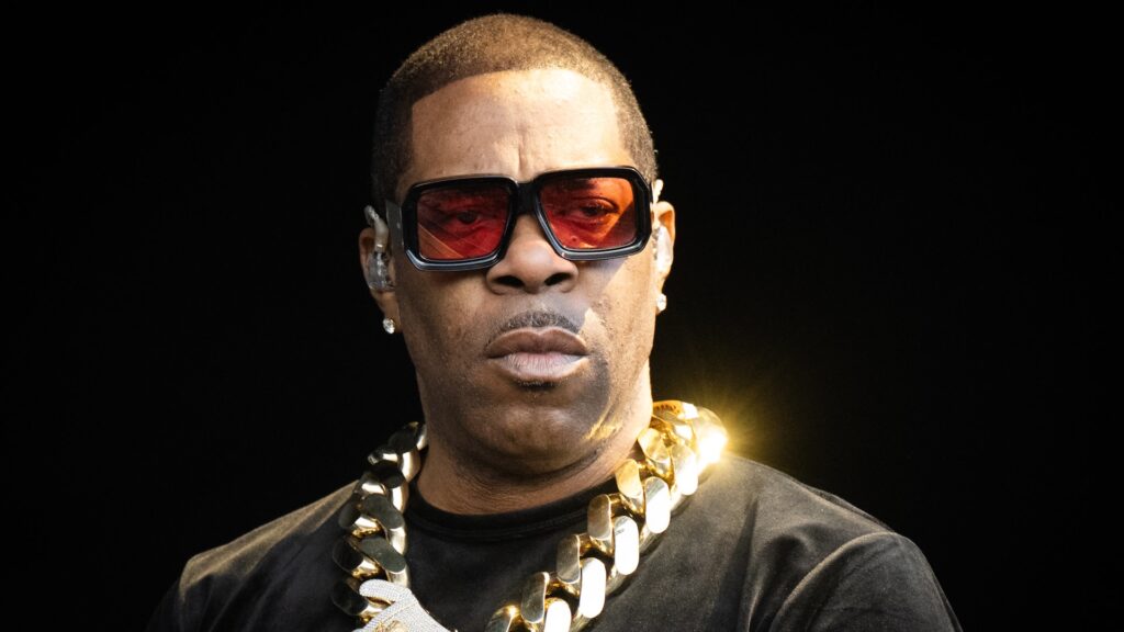 Busta Rhymes wearing large sunglasses and a chunky gold chain, performing on stage.