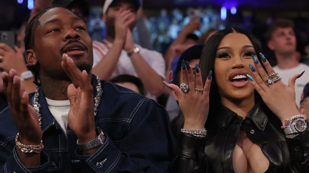A man and a woman sitting in a crowd, clapping and smiling. The woman has long black hair and is showing off her rings.
