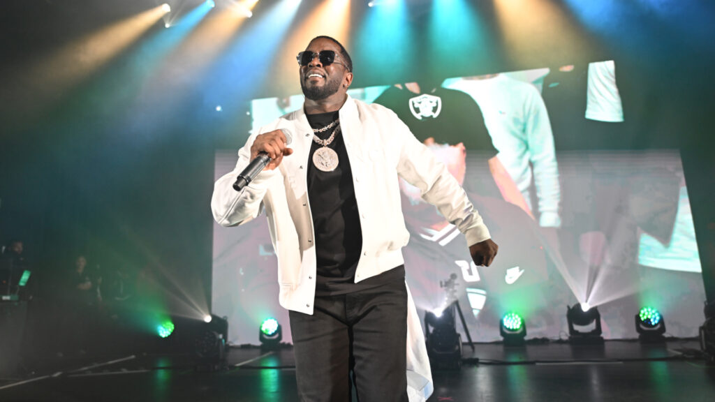 Sean "Diddy" Combs performing on stage, wearing sunglasses and a white jacket, holding a microphone.