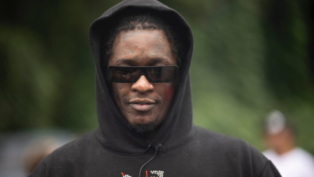 Young Thug in a black hoodie and sunglasses stands outdoors, with a blurred green background.