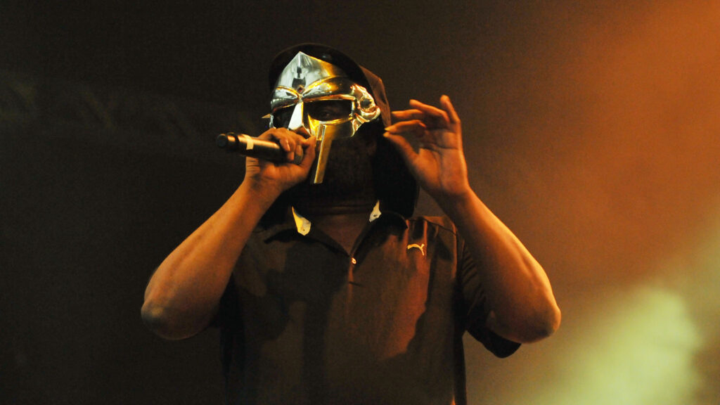 Rapper MF DOOM performing on stage, wearing his iconic metal mask and holding a microphone, with an orange-lit background.