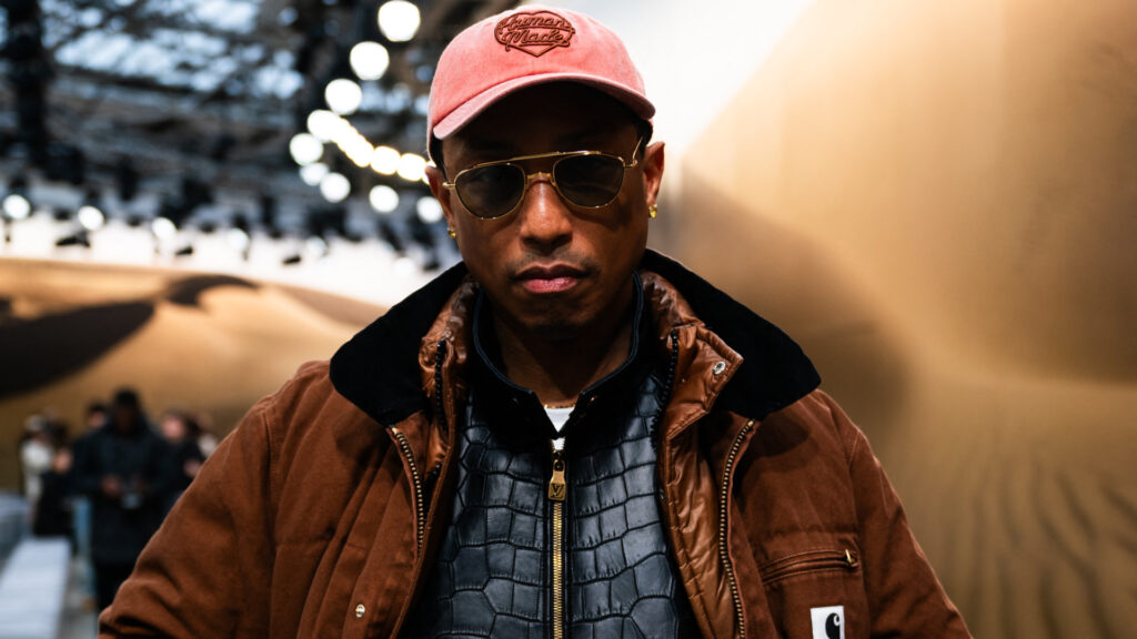 Pharrell Williams wearing sunglasses and a pink cap, dressed in a brown jacket with a patterned shirt underneath at a fashion event.
